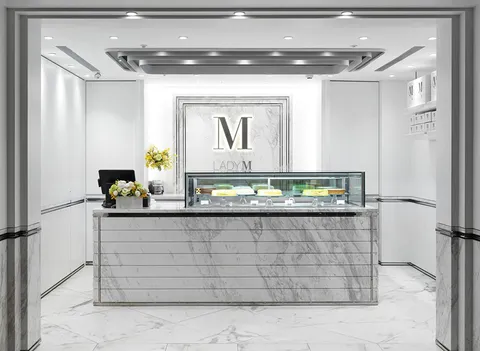 Lady M Regent Taipei: marble counter, cakes, framed logo, recessed lights, yellow flowers. White/gray, elegant.
