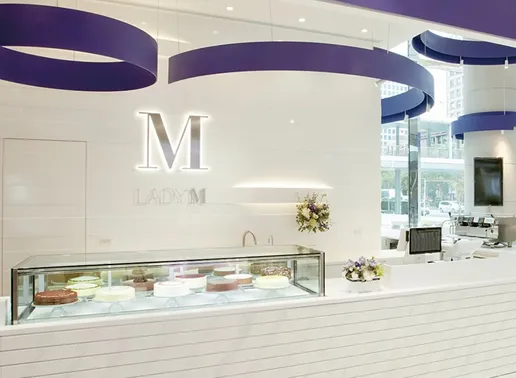 Lady M Far East Xinyi Store Taipei Boutique featuring a white striped counter with a glass display case showing colorful cakes. The space has an illuminated white "M" logo on the wall, distinctive blue circular lighting fixtures suspended from the ceiling, fresh floral arrangements, and large windows providing natural light.
