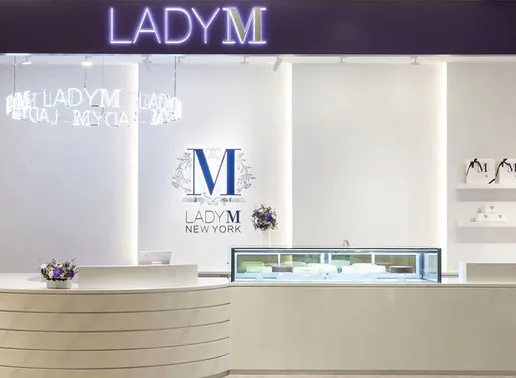 Lady M Mega City Banqiao Store in Taipei featuring white walls with illuminated "LADY M" signage on a navy header. Inside, a circular neon LADY M light fixture hangs above the curved white counter with a glass display case. The blue Lady M logo with decorative elements appears on the back wall, with branded merchandise displayed on shelves and purple floral arrangements adding elegant touches.