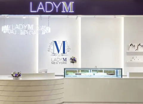 Lady M Banqiao Taipei: lit "LADY M" on navy, neon logo, curved white counter, blue logo, purple flowers, merch.