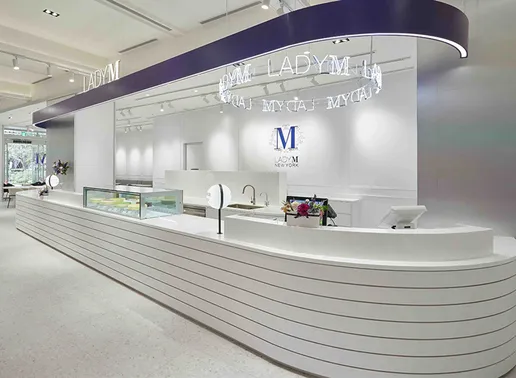 Lady M Taichung Eslite480 Boutique interior featuring a long white striped counter with rounded ends and a glass display case. A circular neon "LADY M" light fixture hangs beneath a navy blue ceiling beam, with the blue Lady M logo on the white back wall.