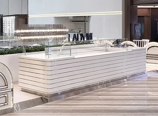 Lady M Jewel Changi Airport Singapore Boutique kiosk with the signature white horizontal striped counter and glass display case. The modern design features a black LADY M sign, crystal lighting fixture overhead, and white chairs visible to the side. The sleek counter sits on a polished marble floor.