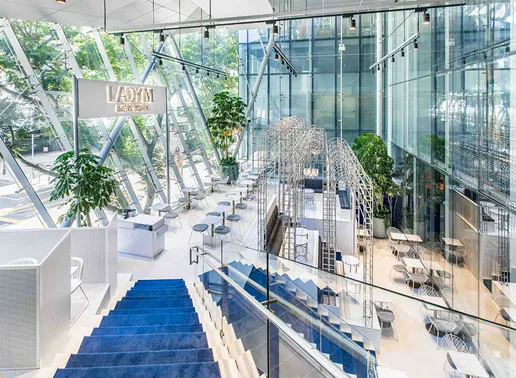 Lady M Orchard Central Singapore Boutique in a glass-walled building with slanted architectural elements and abundant natural light. The space features illuminated LADY M signage, blue stairs, potted plants, white tables with chairs, and a distinctive metal arch installation. The contemporary design creates an airy, sophisticated atmosphere.
