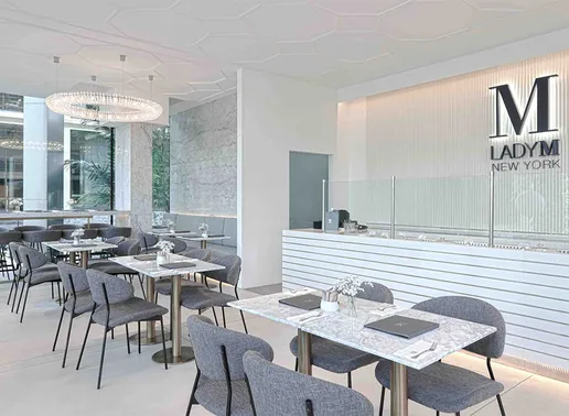 Lady M South Beach Singapore Boutique featuring a sleek dining area with gray upholstered chairs and marble-topped tables. The counter features the signature white horizontal striping with clear glass partitions and the navy blue Lady M logo on the textured white wall. A crystal ring chandelier hangs from the decorative ceiling, creating an elegant, monochromatic atmosphere.