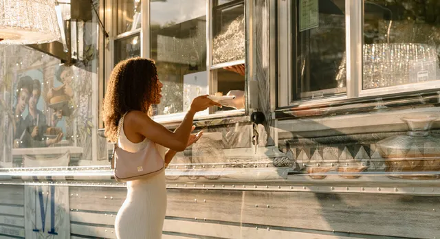 Lady M x Baccarat Luxury Cake Truck guest with curly hair and a white dress ordering cake at truck window in warm sunlight