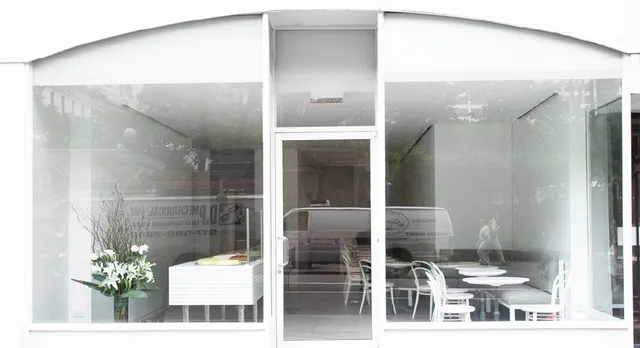 Lady M's Bryant Park storefront with large glass windows and central door. Inside features white tables, chairs, and a cake display case. A simple white flower arrangement sits in the window.