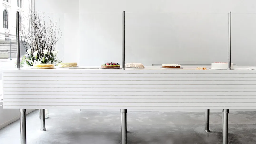 Minimalist cake display at Lady M Bryant Park Boutique featuring a white ribbed counter with chrome legs showcasing an assortment of handmade cakes behind glass partitions. A decorative arrangement of white flowers and curly branches sits at one end. The space has concrete flooring and a bright, clean aesthetic with a historical building visible through the window.