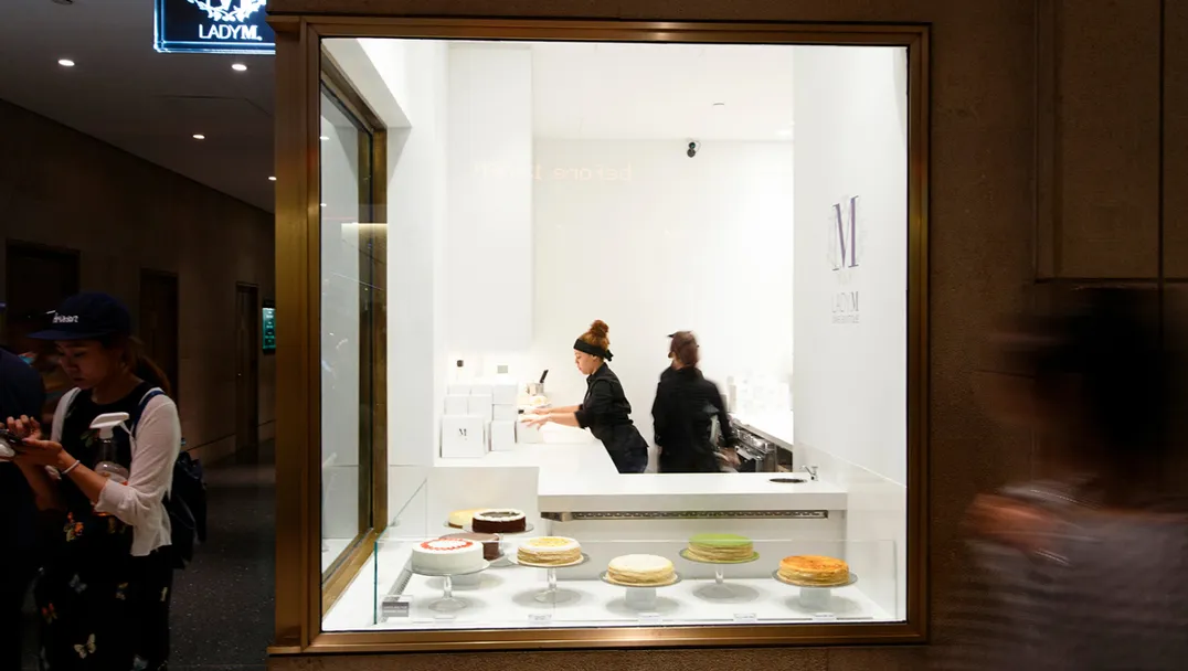 Lady M Rockefeller Center Boutique storefront with illuminated logo sign. Display window showcases various cakes on white pedestal cake stands. Staff in black uniforms work inside the bright white interior and cake boxes with the 'M' logo are visible on shelves behind the counter.