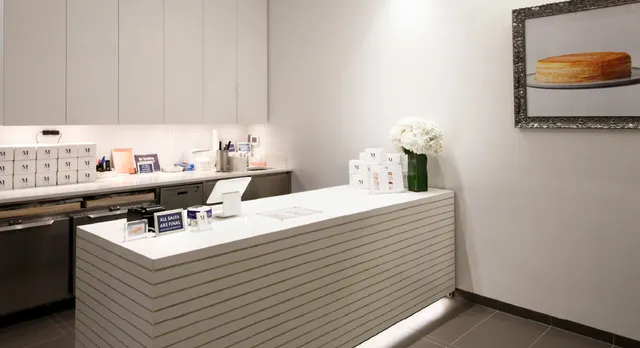 Interior of a Lady M Rockefeller Center featuring a minimalist design with white and gray tones. The space includes a white ribbed front counter with a point-of-sale system and menu displays. On the wall hangs a framed photograph of a Signature Mille Crêpes cake, and a small white floral arrangement adds a touch of elegance to the counter.