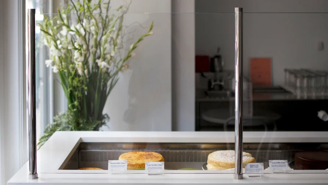 Lady M NoMad minimal cake display case with several whole cakes behind glass and small description cards. Behind the display features an elegant arrangement of green stems and white flowers on the left side.