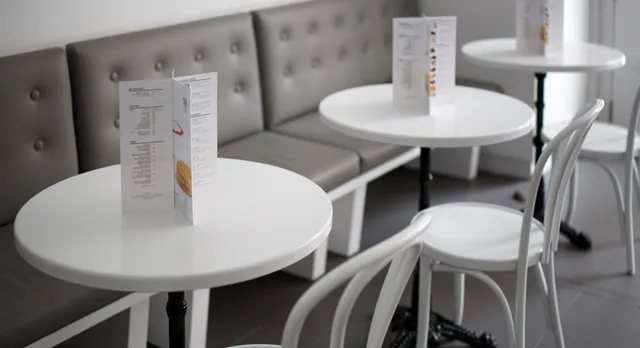 The interior of Lady M's NoMad Boutique, featuring three small, round white tables with black bases. Each table has a white chair pulled up to it and a menu standing upright. A long, tufted gray bench lines the wall behind the tables.