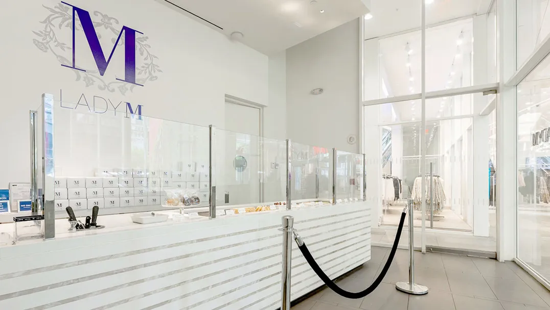 Bright, modern interior of Lady M World Trade Center Boutique featuring a long white cake display with horizontal marble detail and clear glass partitions. A large purple "M" logo with floral accents and "LADY M" in smaller letters is on the white wall above the cakes. The floor is light gray tile, and glimpses of a brightly lit storefront are visible in the background.