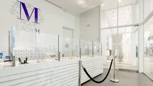 Bright, modern Lady M WTC: long white marble cake display, purple floral "M" logo, light gray tile.