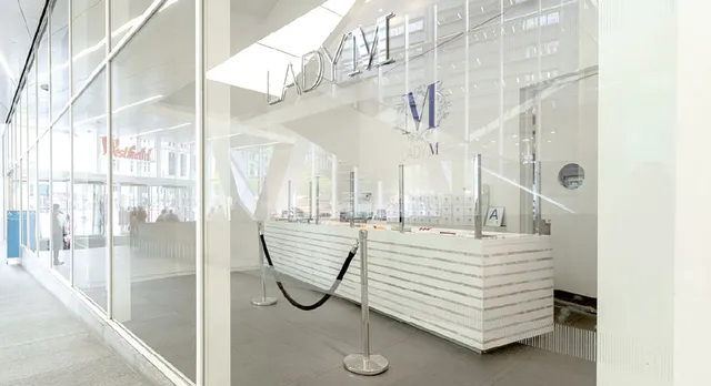 A brightly lit storefront of Lady M World Trade Center is visible through large glass windows. Inside, a white counter with horizontal marble accents and clear glass partitions is visible displaying a selection of cakes. The interior is white and brightly illuminated. Outside, a glimpse of the Westfield shopping mall walkway and people can be seen.