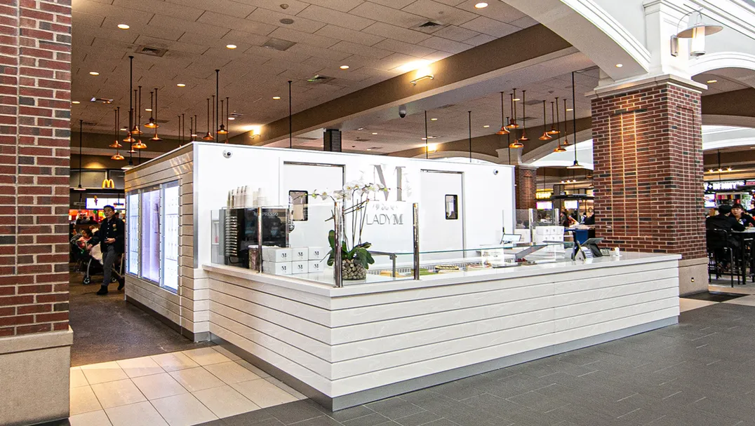 A brightly lit Lady M Cake Boutique kiosk situated in the Woodbury Common Food Court. The kiosk has a white cake display with glass showcasing cakes, and a white backdrop with the purple "LADYM" logo.