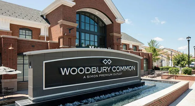 Entrance sign for "Woodbury Common Premium Outlet" in front of a brick outlet mall on a sunny day. Water feature in the foreground.