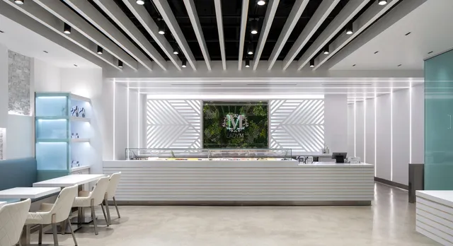 Interior of Lady M Sawtelle featuring a long, white cake display. Behind the counter is a floral "M" logo with greenery. Ceiling features black and white parallel beams. Small white tables and light-colored chairs are arranged on the left side of the space.
