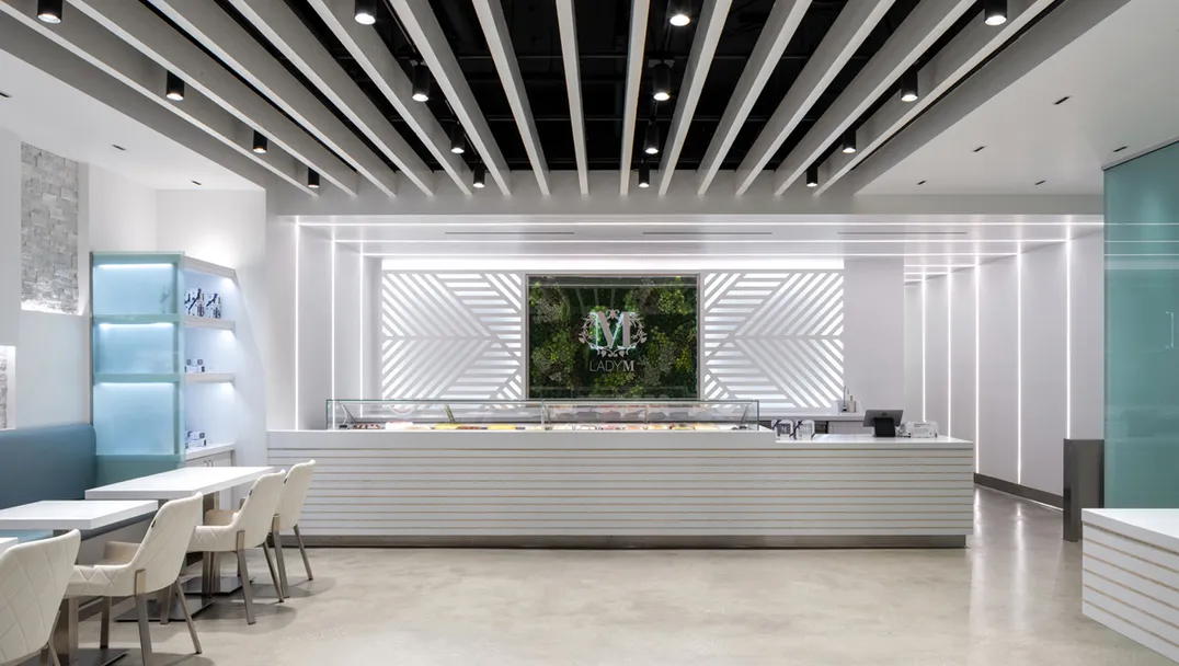 Interior of Lady M Sawtelle featuring a long, white cake display. Behind the counter is a floral "M" logo with greenery. Ceiling features black and white parallel beams. Small white tables and light-colored chairs are arranged on the left side of the space.