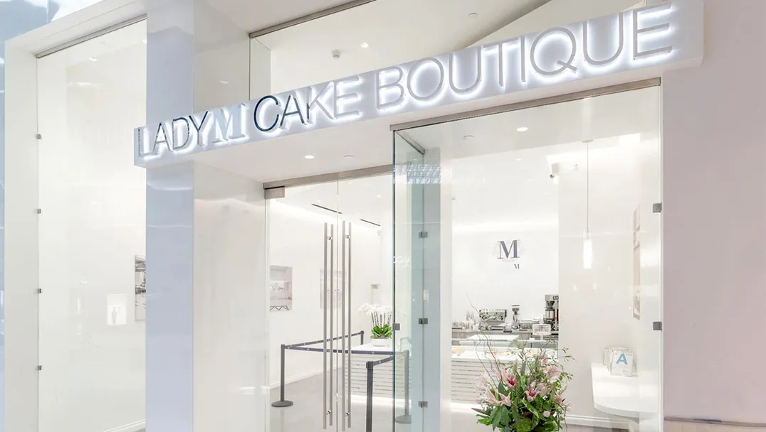 Storefront of LADY M CAKE BOUTIQUE in Arcadia, CA within The Shops at Santa Anita featuring illuminated signage, glass doors, bright white interior, and a floral arrangement by the entrance. The design is modern and elegant with minimalist aesthetic.