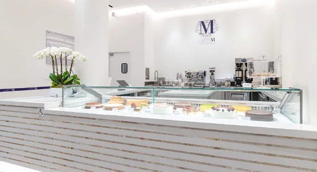 Interior of LADY M boutique in Arcadia, CA featuring a pristine white display counter with glass pastry case showing assorted cakes and desserts. White orchids decorate the counter, and the LADY M logo is visible on the back wall above coffee equipment.