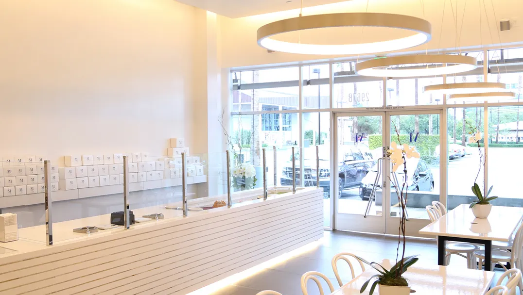 Lady M Irvine boutique featuring a white counter with a cake display, circular pendant lighting, and large glass windows that let in natural light. Small tables with white chairs are arranged for seating, and potted orchids add subtle decoration to the sleek environment.