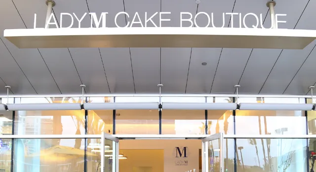 LADY M CAKE BOUTIQUE storefront entrance sign above glass doors in Irvine, CA. Through the glass doors, the brand's "M" logo is visible inside the boutique.