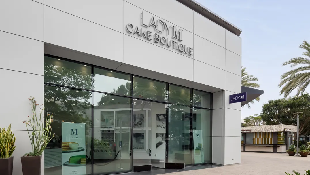Modern white Lady M San Diego storefront with large glass windows and silver signage. The entrance features glass doors and display windows showing "M" branding inside. Decorative plants are positioned near the entrance, with palm trees visible to the right. The contemporary design sits on a paved walkway.