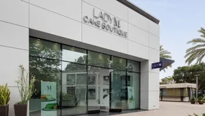 Modern white Lady M San Diego storefront with glass windows, silver sign, plants, and palm trees.