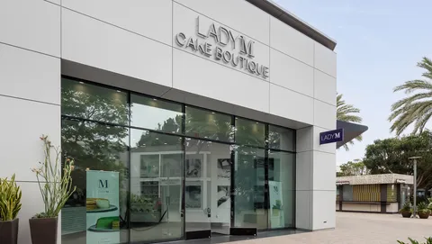 Modern white Lady M San Diego storefront with glass windows, silver sign, plants, and palm trees.