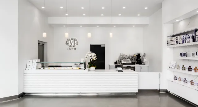 Interior of Lady M San Diego featuring a minimalist white design. A sleek white counter with horizontal ridges and glass display case showcases cakes. The LADY M logo appears on the back wall above coffee equipment. Pendant lights hang from the ceiling, and retail products are displayed on wall shelves to the right. An orchid plant decorates the counter.