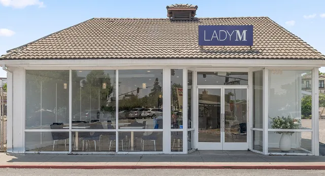 A small storefront with "LADY M" signage on a tiled roof. The building features large glass windows and doors, with indoor seating visible through the glass. A white planter sits inside at the entrance.