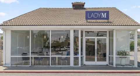 Small storefront with "LADY M" sign on tiled roof, large glass windows/doors, visible indoor seating, white entrance planter.
