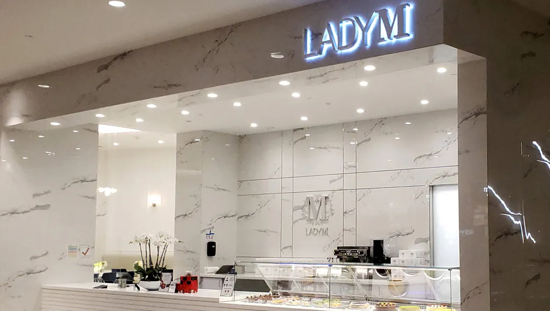 An elegant Lady M counter located at Westfield Valley Fair with a sleek design featuring white marble walls and surfaces. The illuminated "LADY M" sign glows above the service area. The display case showcases colorful cakes, while a small white orchid decorates the counter. The minimalist space is brightly lit with recessed lighting.