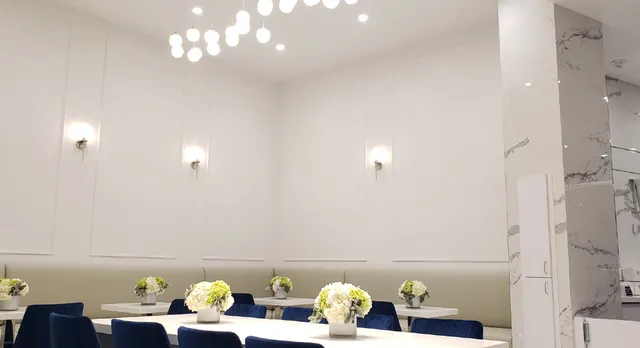 A bright, elegant dining area with white walls accented by wall sconces. Navy blue velvet chairs surround white tables, each adorned with small arrangements of white and light green flowers. Hanging pendant lights with round bulbs illuminate the space from above, and a marble column is visible on the right side.