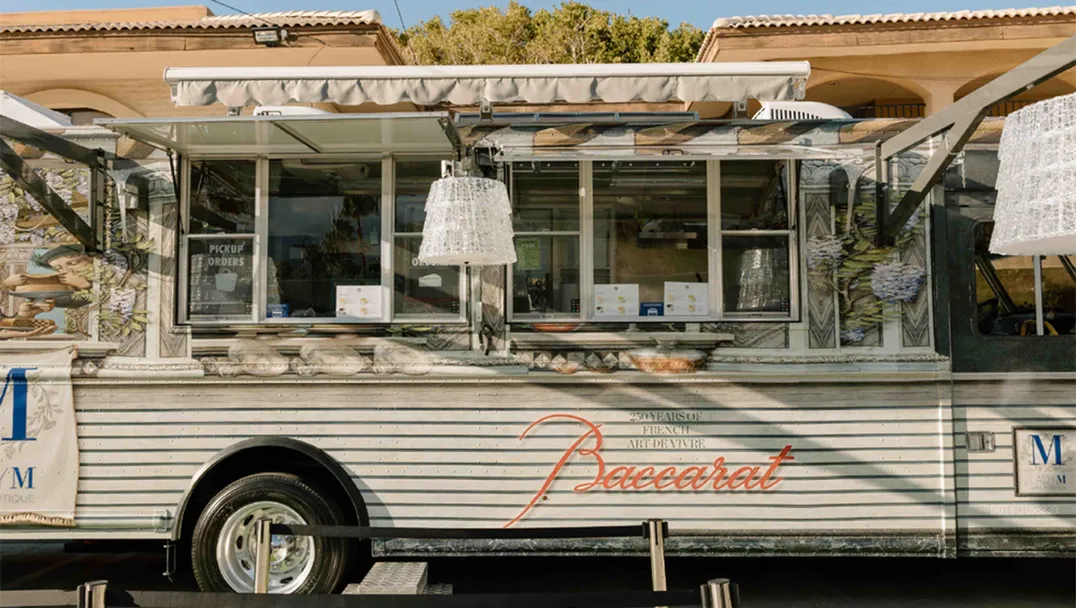 A luxury cake truck with "Baccarat" and "LADY M" logos on its side. The silver trailer features a retractable awning, serving windows with pendant lamps, Baccarat chandeliers and is parked on a green surface.