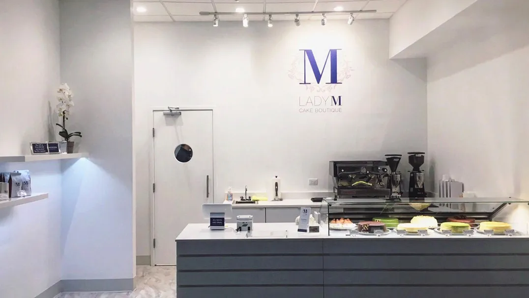 Lady M Chicago boutique with white walls featuring the Lady M logo. The space has a modern gray counter with a glass display case showing various cakes. A coffee machine sits on the counter, and a white orchid decorates the space.