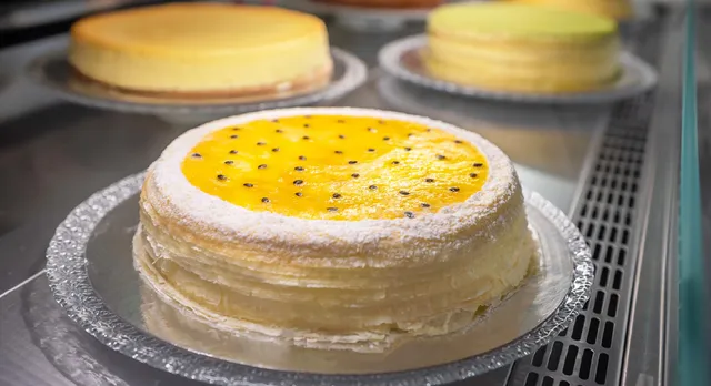 A Passion Fruit Mille Crêpes cake with a passion fruit glaze topping and passion fruit seeds and visible layers of thin crepes. Signature and Green Tea cakes are visible in the background on a refrigerated display shelf.