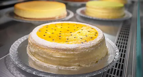 Passion Fruit Mille Crêpes cake with glaze, seeds, and visible thin layers. Other cakes behind.