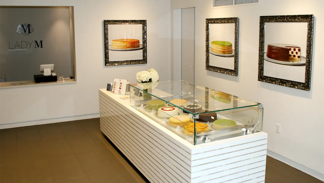 Interior of a Lady M Boston featuring a white display counter with glass case containing cakes. On the walls are framed photographs of signature cakes, and a Lady M logo is visible on the left wall.