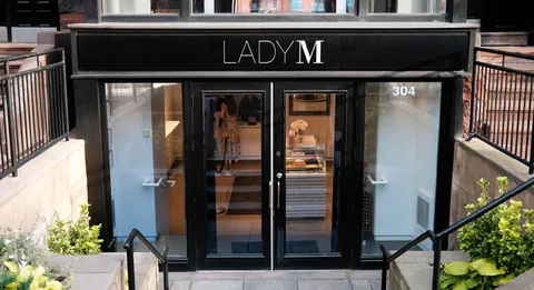 Lady M Boston storefront with black frame, glass doors, logo on awning, and steps with planters.