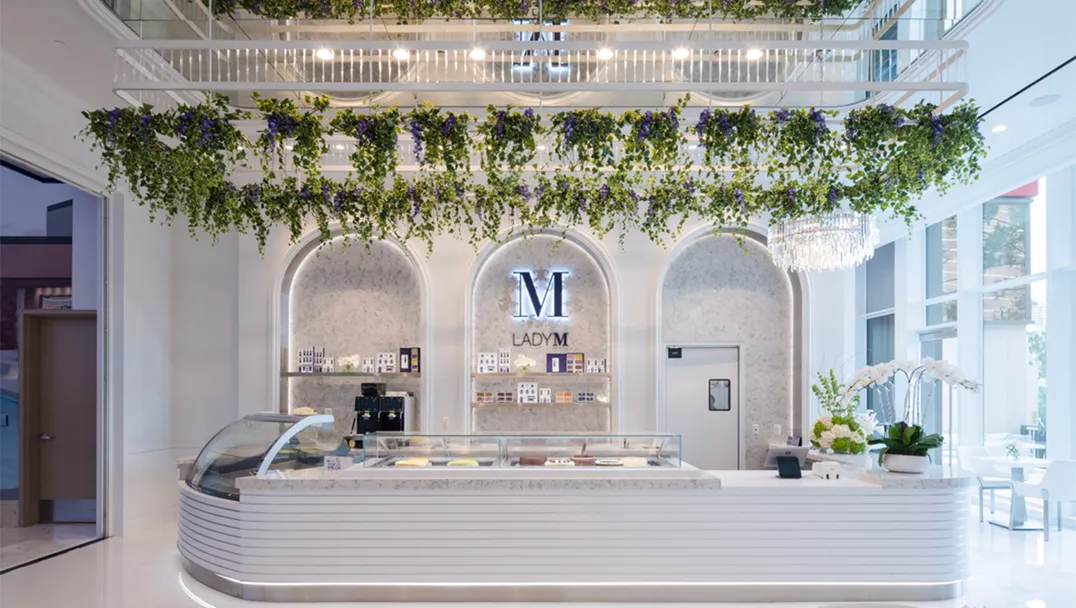 Interior of Lady M at Resorts World Las Vegas featuring an elegant white cake display counter with glass display case. Behind the counter are three arched alcoves with marble backing and the Lady M logo. Hanging greenery and plants create a garden-like canopy above the counter, and a crystal chandelier adds a touch of luxury to the bright, airy space.