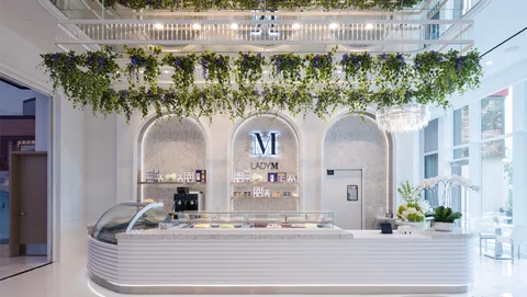 Lady M Resorts World Vegas interior with white cake counter, arched alcoves, greenery, and chandelier.