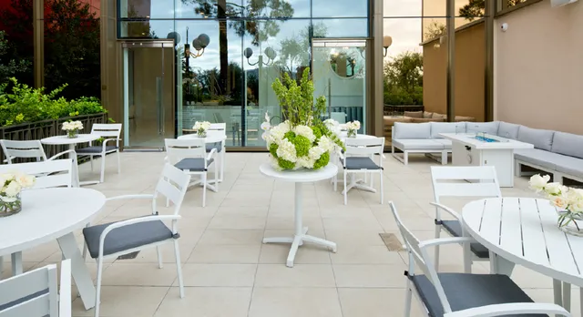Outdoor seating area at Lady M Las Vegas featuring white tables and chairs with gray cushions arranged on a light stone patio. A central table displays a fresh floral arrangement with white and green flowers. Large glass windows reflect surrounding greenery, and comfortable lounge seating is visible on the right side.