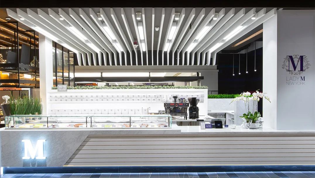 Lady M's modern and stylish dessert kiosk at American Dream featuring a sleek white design with clean lines and slatted paneling. The counter showcases an illuminated "M" logo, and a glass display case is filled with an assortment of colorful cakes. Overhead, geometric ceiling panels with integrated lighting add a contemporary touch, while fresh greenery and orchids enhance the inviting atmosphere.