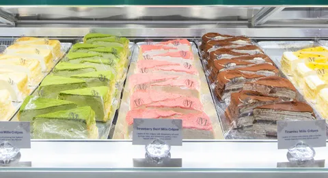 Close-up of Mille Crêpes slices in display case with "M" logo and flavor labels.