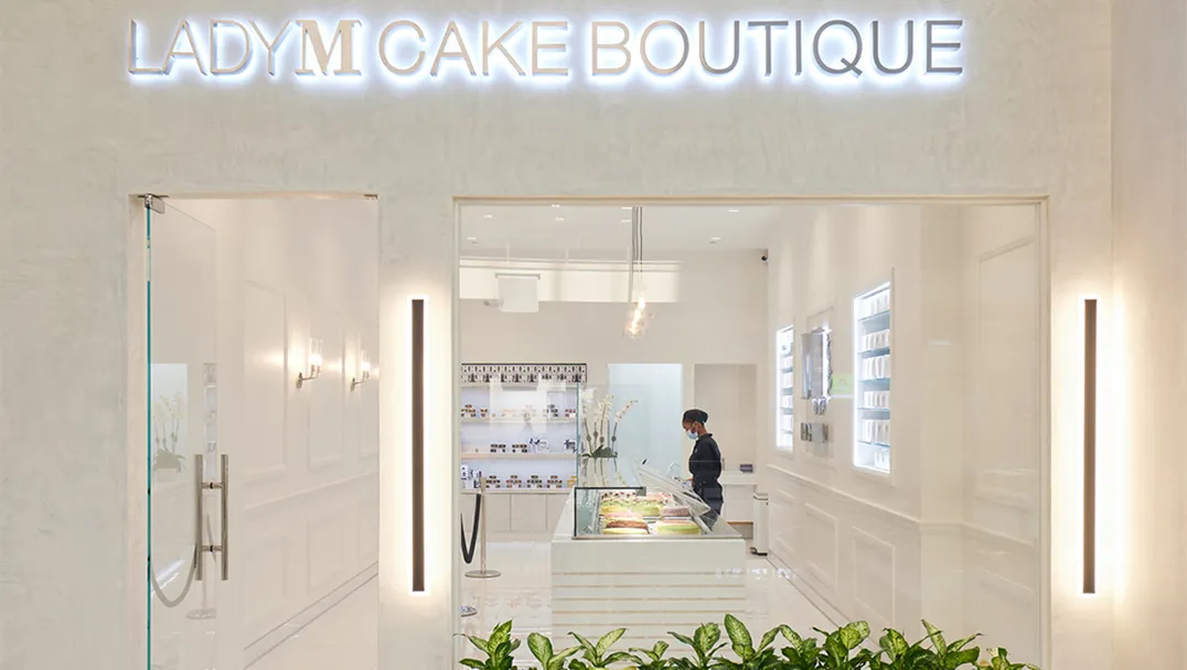 Front entrance of Lady M Cake Boutique with illuminated signage, a modern white interior, display case with cakes, and a person behind the counter.