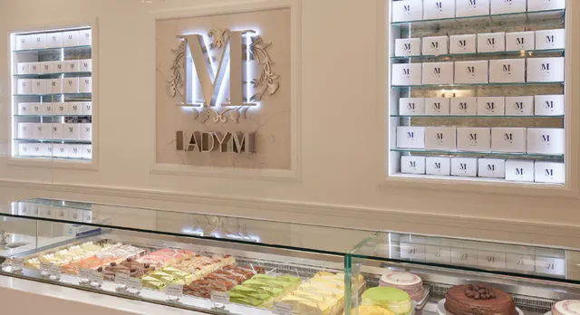 Interior of Lady M Cake Boutique at The Galleria in Houston showing a display case filled with colorful slices of Mille Crêpes cakes, illuminated "M" logo on the wall, and shelves filled with small and elegant cake boxes.