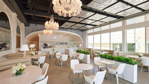 Lady M Houston interior with chandeliers, natural light, white tables, and arched cake counter.