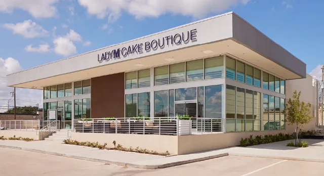 The modern exterior of Lady M Cake Boutique at Houston Westchase featuring a sleek white building with large glass windows, the Lady M logo prominently displayed across the top, and an outdoor patio area with tables and planters.