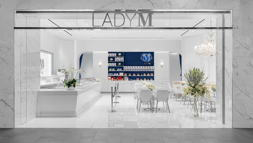 Elegant storefront of Lady M at Tysons Galleria featuring a marble exterior with the LADY M logo prominently displayed above the entrance. Inside, a pristine white interior showcases a cake display counter on the left, a blue accent wall with product displays in the back, and several small white tables and chairs for guests to enjoy their desserts.